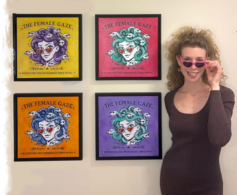 Lauren Farrow in heart-shaped pink sunglasses standing beside her four framed Female Gaze Medusa paintings in yellow, magenta, orange and purple